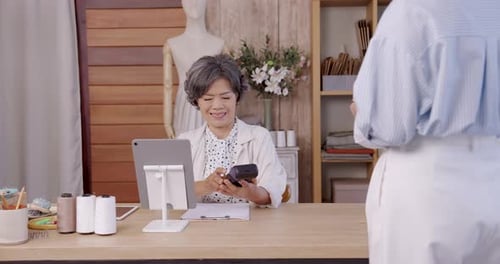 Asian senior tailor processes cashless credit card payment using POS terminal while customer