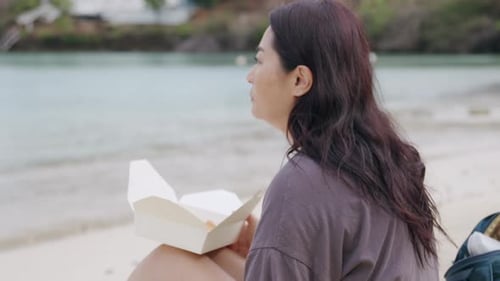 Asian woman eating takeout food while sitting on a beach, portrait mid shot