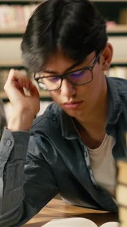 Vertical Video Tired Asian Student Leaning Over Open Book Between Tall Stacks in Library