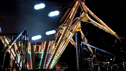 A handheld shot captures an amusement park attraction with people at night, adorned with lights.