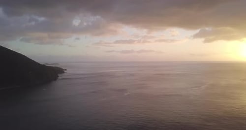 Aerial, flying high over the ocean at sunset. Green mountain and an island in the distance. View of