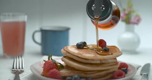Maple Syrup Is Poured Over A Stack Of Pancakes With Fruit And Butter On A Table With A Background...