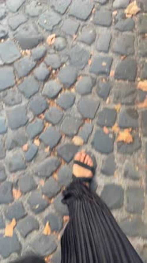 POV shot of female feet with sandals walking on sidewalk or road with cobble stone and leaves on