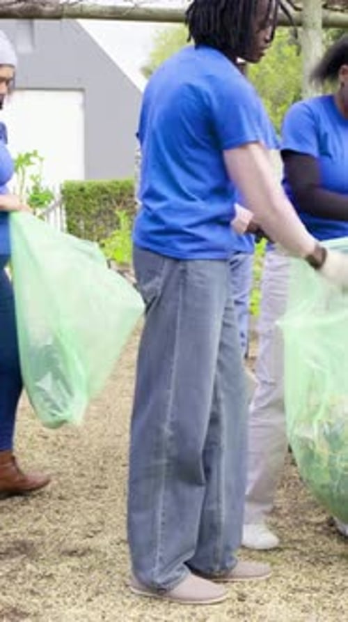 Vídeo vertical: Con una bolsa verde abierta, una mujer vestida con una camiseta azul recolectando esquejes en el jardín para limpiarlos