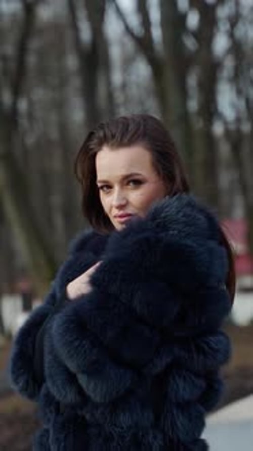 Young beautiful fashionable lady wearing stylish dark fur coat posing in park. Model looking aside.