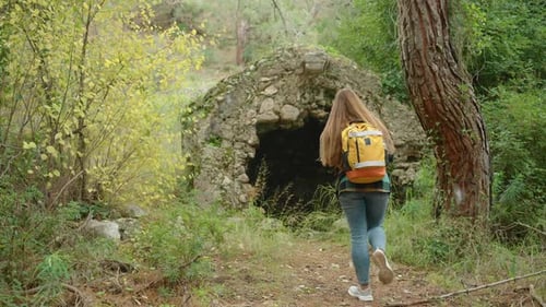 Una mujer recorre el bosque con estructuras antiguas y se asoma a una cripta de piedra.