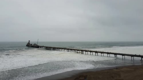 Drone video of a pier in the ocean shore. Drone orbits around while waves crash into the walkway of