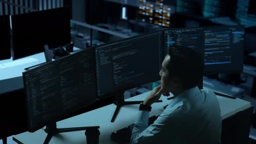 Hombre asiático pensando en algo mientras escribe código con múltiples pantallas de ordenador en la oficina