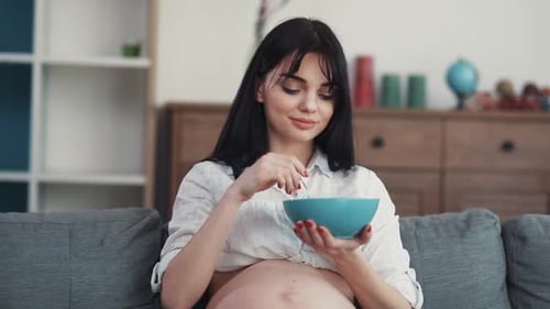 Pregnant woman eating from bowl on grey sofa
