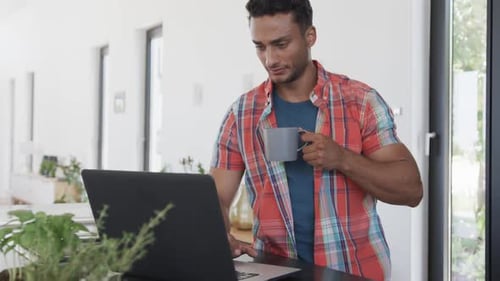 Biracial man having coffee and using laptop in sunny modern apartment, slow motion