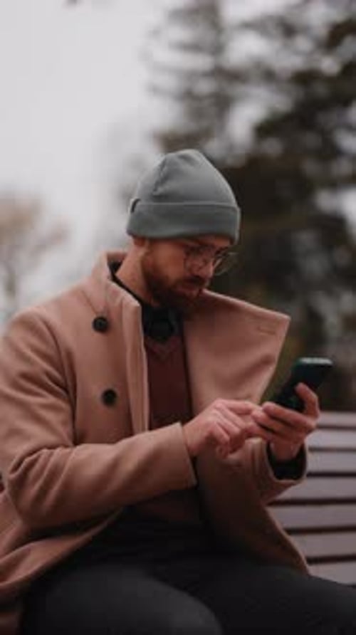 Young Man in a Warm Coat and Beanie Uses His Mobile Phone While Sitting on a Park Bench