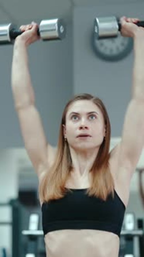 Pretty woman in black top raises up dumbbells on the background of the gym. Exercises for biceps.
