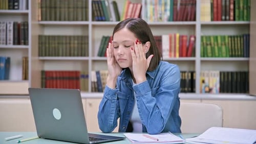 Tired Young University Student in Library Touching Head with Hands