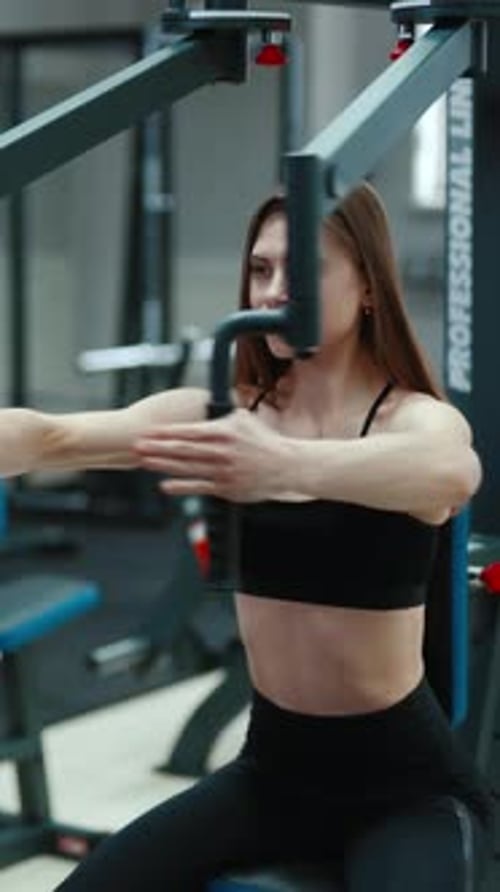 A pretty woman with flowing hair is sitting and doing exercise for the chest in the gym.