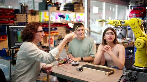 Junge glückliche Ingenieure programmieren in der Werkstatt einen gelben Roboterarm, um Wasserglas aus Pappe zu schnappen