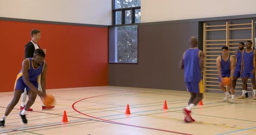 Basketball players practicing dribbling skills in gym with coach encouraging