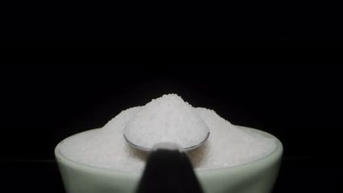 A full spoon of sugar moves backward, moving away from the sugar bowl. Dolly on a black background.