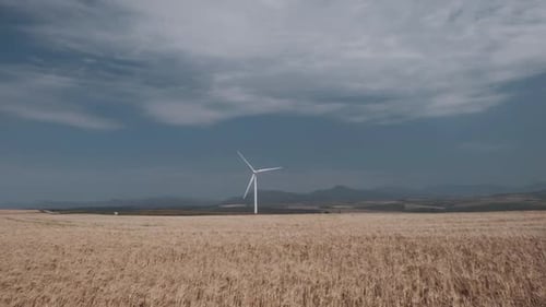 A wind turbine rotating on a wheat field with blue skies clouds and mountains