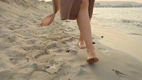 Closeup of female bare feet in long skirt walking on the sandy sea beach at sunset