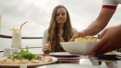 The waiter brings the young woman plates with food, she rubs her hands. An open-air restaurant high