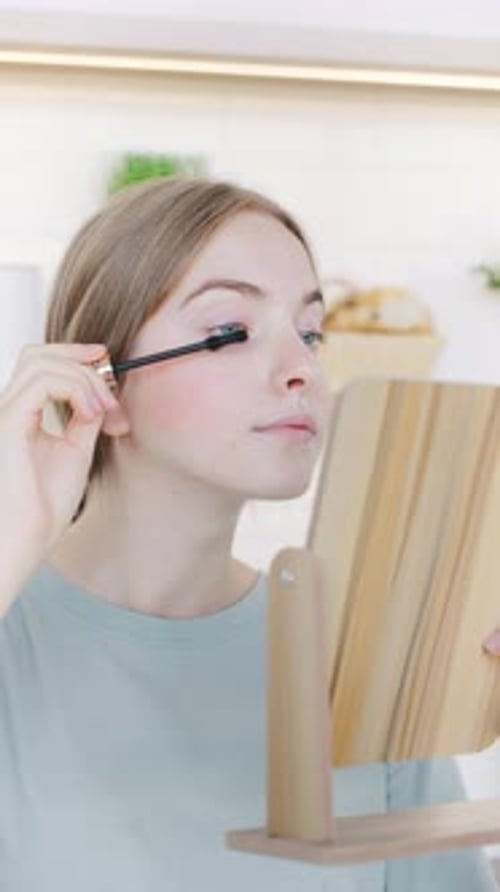 Young Woman Doing Makeup Applies Mascara While Looking Into a Wooden Mirror in a Bright Modern