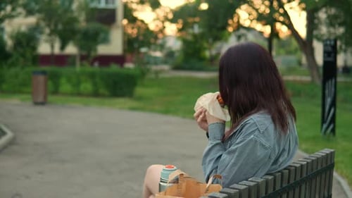 Back View of Young Adult Woman in Casual Clothes Having Break in the Park and Eating Hamburger