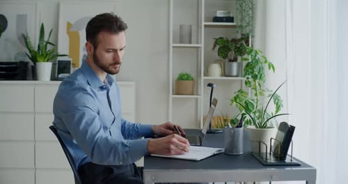 Man Writing at Desk with Laptop at Home