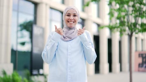 Young smiling muslim female employee in hijab showing thumb up looking at camera standing on street