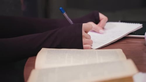 Close Up Of Woman Writing In Notebook With Blurred Book On Brown Table