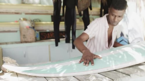 African american man preparing surfboard on the counter of surf rental beach shack, slow motion