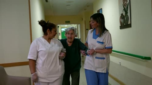 Nurses Assisting Elderly Woman in Walking Down Nursing Home Corridor