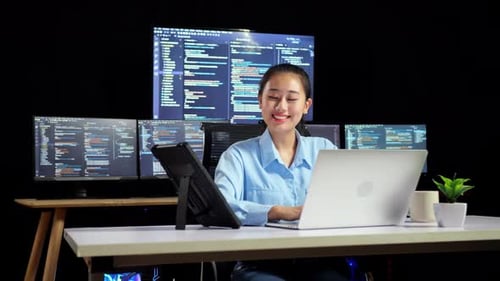 Programadora asiática mirando una tableta y escribiendo código con una computadora portátil usando varios monitores