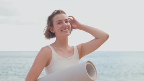 Smiling Young Woman Holding Yoga Mat By Ocean at Beach