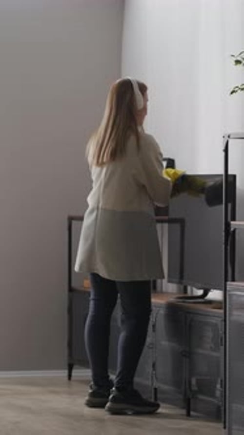 Young Woman Wearing Headphones and Rubber Gloves Dusting Shelves and Furniture in Modern Minimalist