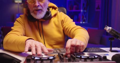 Close Up of Cool Stylish Caucasian Bearded Senior Man DJ Sitting at Desk in Dark Room with Neon