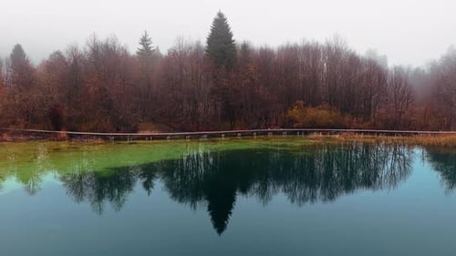 Drohnenaufnahme eines ruhigen Sees, umgeben von dichtem Herbstwald unter einer Schicht Morgennebel in P