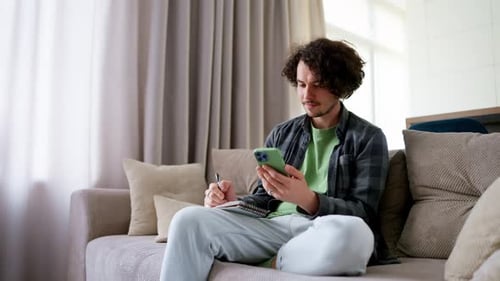 Young Adult Writing in Notebook on Sofa Using Phone