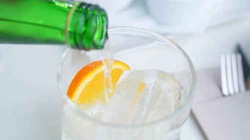 Pouring soda into glass with ice and slice of orange, close up