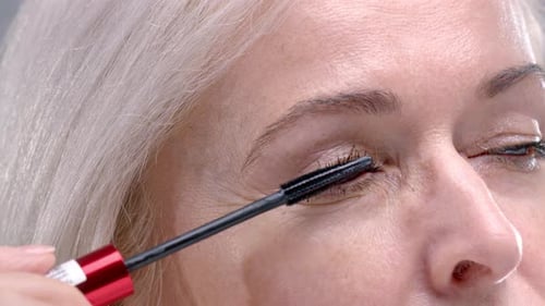 Blonde Senior Lady with Gray Eyes Applies Mascara with Professional Brush on Light Grey Background
