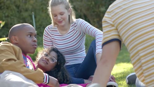Happy diverse group of teenage friends sitting on grass and talking in sunny park, slow motion