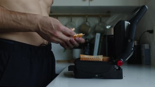 Man walks up to a panini grill press and places marble cheese slices onto two pieces of white bread,