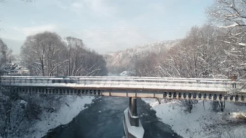 Aerial view of car crossing the bridge covered by snow, frozen river and snowy forest.