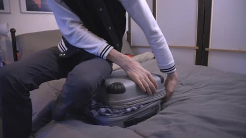 A Young Man Sits On His Overloaded Travel Suitcase Trying To Close With Both Hands - Half-Body Shot
