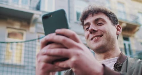 Caucasian Cheerful Young Man Standing at Street and Texting Message on Mobile Phone Outside Handsome