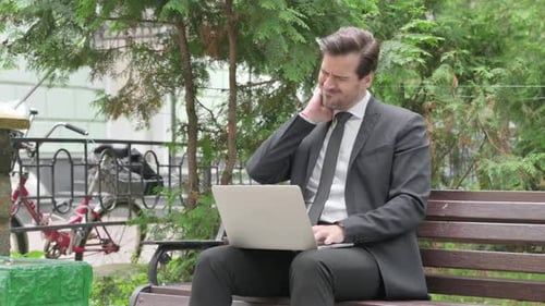 Businessman Types on Laptop and Stretches Neck in Park