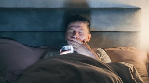 Young man in bed at home late at night scrolling mobile phone in low light and yawn