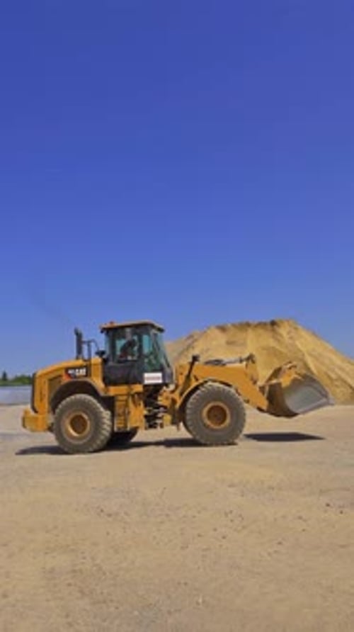 Excavadora moviéndose entre montones de grava. Planta de asfalto. Maquinaria industrial que trabaja al aire libre.