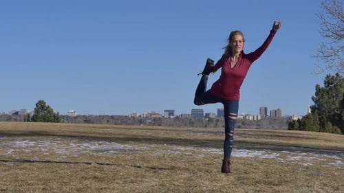 Blonde woman doing Dancer’s Pose (Natarajasana) on yellow grass in the park in the winter