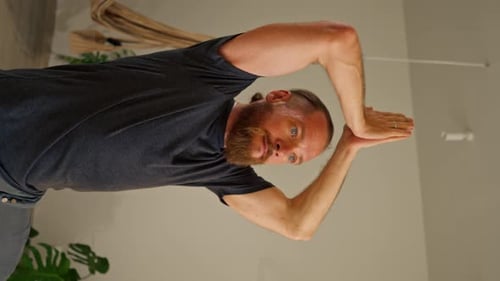 vertical video an adult man with a beard and long hair stands on one leg in the hall doing yoga