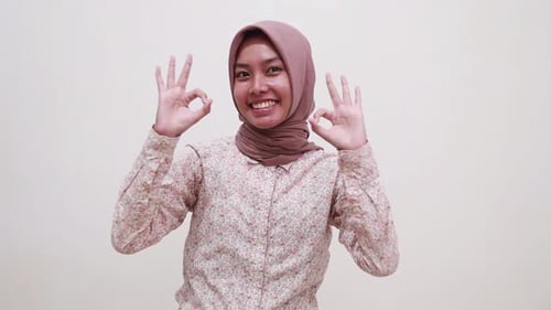 Excited young asian muslim woman standing pointing down and showing okay hand gesture. Isolated on w
